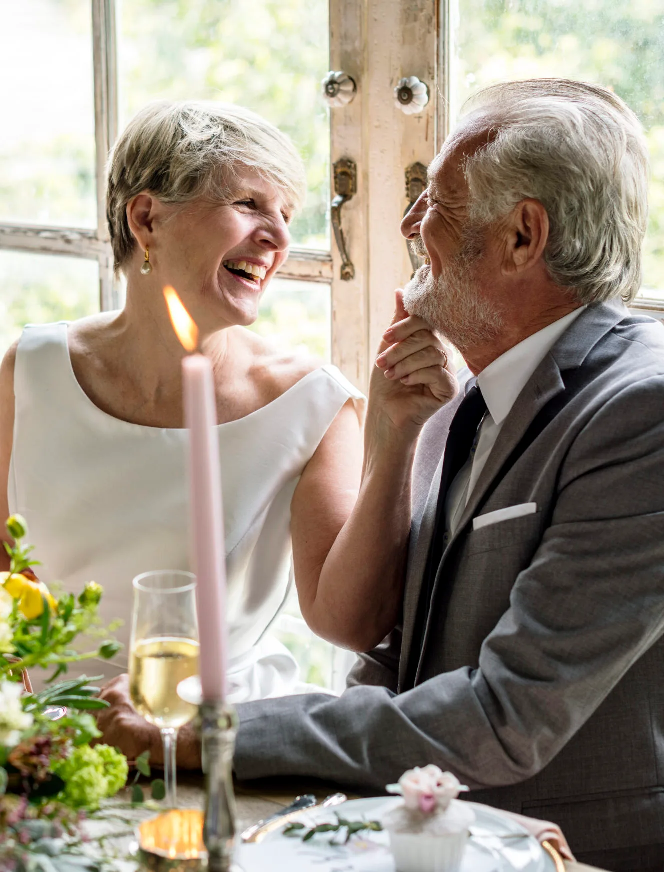 A man and woman dressed for a wedding smiling at each other whilst sitting at a table with a candle.