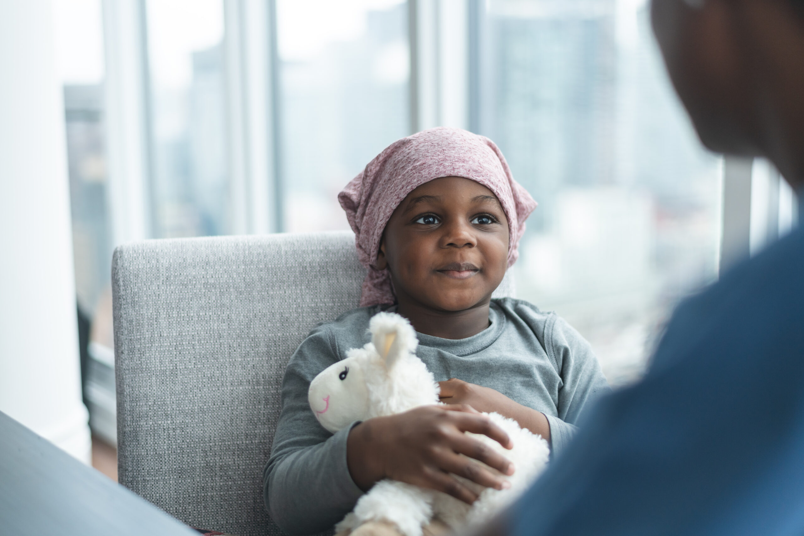 A young boy of African descent is at a medical consultation. He is dressed in casual clothes while wearing a headscarf to hide his hair loss. He hugs his stuffed animal while listening to his doctor of African descent comfort him.